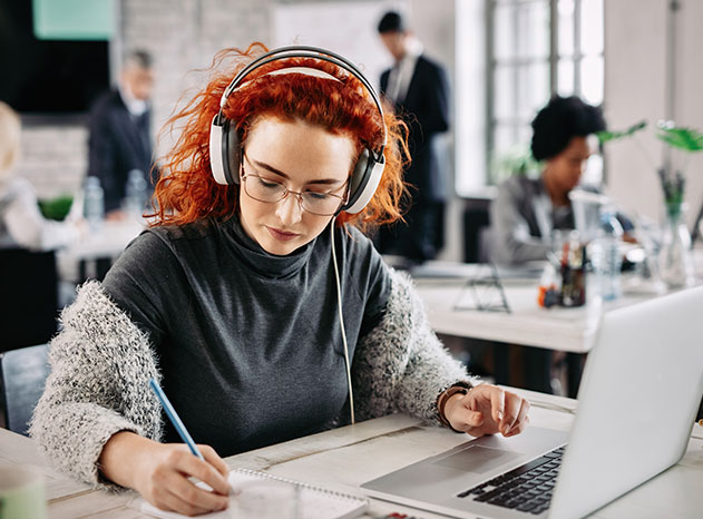 Dona amb auriculars escriu en una llibreta davant d'un portàtil en una oficina